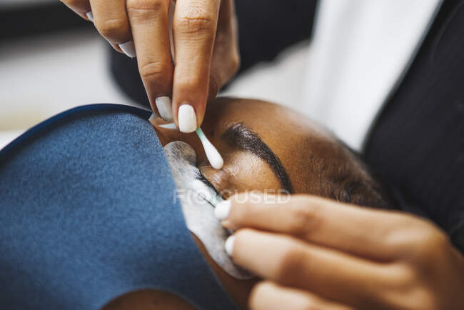 Crop unrecognizable beauty technician with cotton bud preparing ethnic woman with patch for eyelash extension procedure in salon — Foto stock