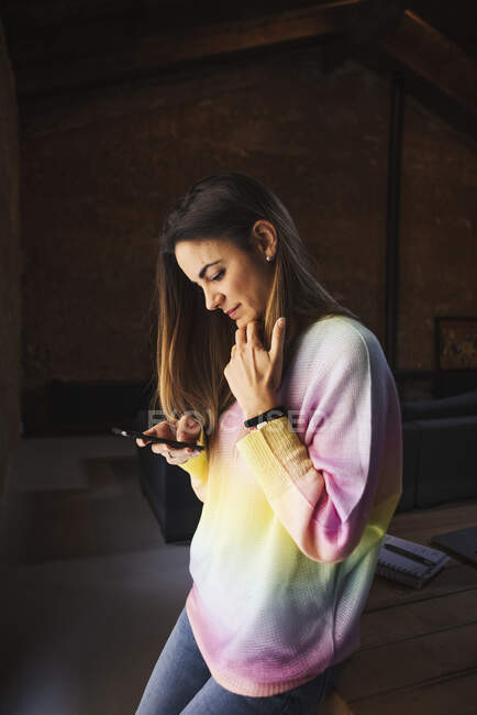Вид збоку на концентрований жіночий серфінг Інтернет на мобільному телефоні, сидячи на столі в будинку в стилі лофт — стокове фото