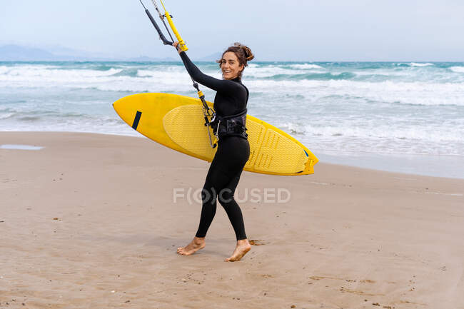 Sportswoman in wetsuit with inflatable kite strolling on sandy shore while looking at camera against stormy ocean — Stock Photo