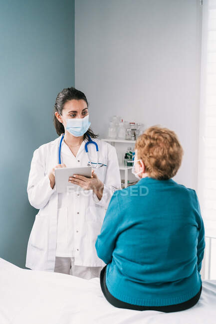 Жінка-лікар у формі з таблетками, що розмовляють зі старшою жінкою у стерильній масці за консультацією, дивлячись один на одного під час пандемії коронавірусу — стокове фото