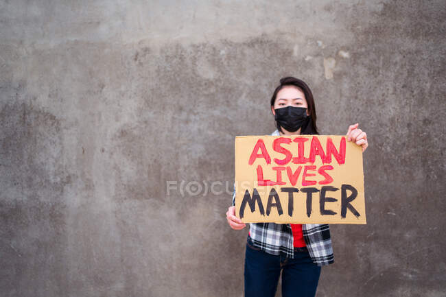 Этническая женщина в маске и с картонным плакатом с надписью Asian Lives Matter protesting in city street and looking at camera — стоковое фото
