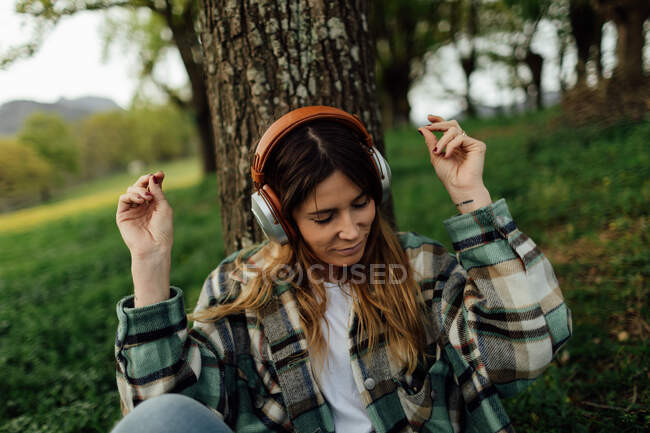 Young content female with tattoo in checkered shirt listening to music from headphones on lawn in summer — Photo de stock