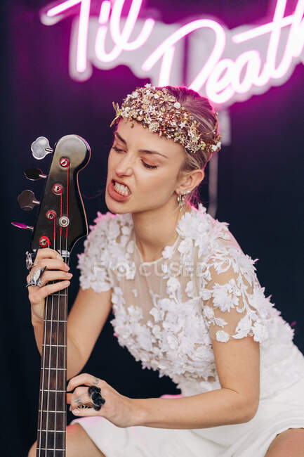 Energetic rebellious young woman in elegant white bridal dress and wreath with guitar in hand making faces in studio with neon inscription — Fotografia de Stock