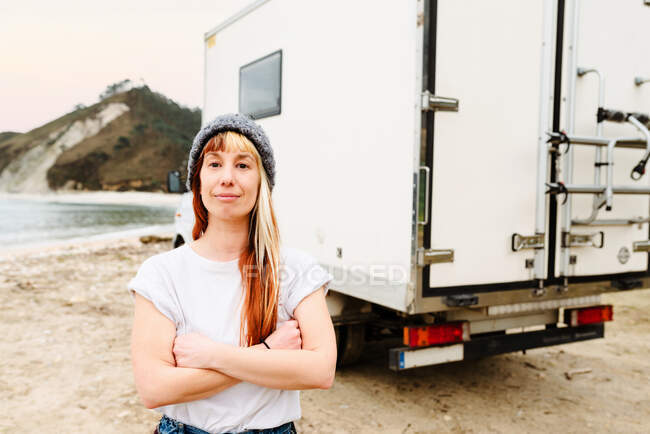 Позитивна жінка-мандрівник стоїть з схрещеними руками біля вантажівки, припаркованої на березі озера в горах і дивлячись на камеру — стокове фото