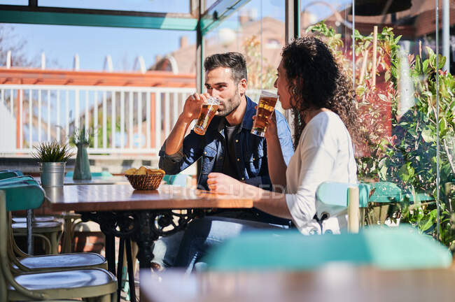 Alegre pareja étnica bebiendo cerveza con papas fritas en un restaurante a la luz del sol - foto de stock