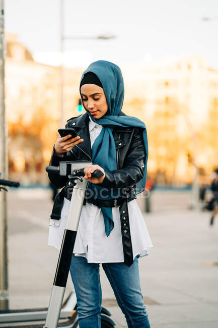 Contenido Mujer musulmana en el pañuelo tradicional usando la aplicación en el teléfono inteligente y la pantalla de escaneo para desbloquear scooter eléctrico moderno estacionado en la calle de la ciudad - foto de stock