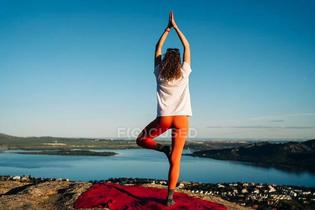 Back view of anonymous female traveler in sportswear standing in Tree with Arms Up asana while practicing yoga on rocky mountain over seacoast against cloudless blue sky — Stock Photo