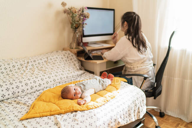 Maman anonyme contre ordinateur de bureau et petit enfant couché sur un lit doux dans la pièce de la maison légère — Photo de stock