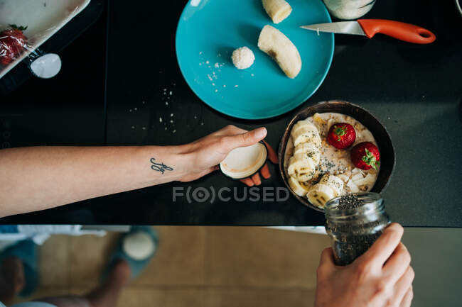Top view crop unrecognizable person with black sesame topping tasty healthy porridge with banana and strawberries in kitchen — Photo de stock