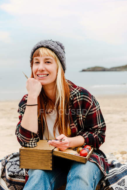 Lächelnde Kunsthandwerkerin, die handgefertigte Bijouterie herstellt, während sie Halbedelsteine verwendet und am Strand am Meer sitzt und in die Kamera schaut — Stockfoto