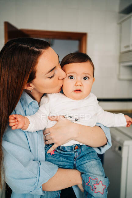 Vista laterale giovane madre in abiti casual che abbraccia e bacia il piccolo bambino nella stanza della luce — Foto stock