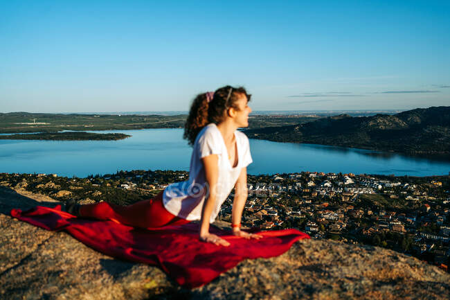 Vista lateral de la joven viajera feliz en ropa deportiva sonriendo mientras practica Upward Facing Dog con cuello Stretch yoga asana en acantilado rocoso bajo cielo azul sin nubes - foto de stock