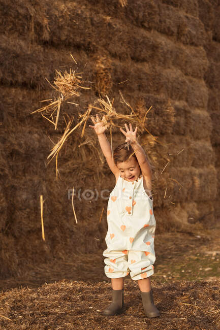 Criança adorável alegre em macacões brincando com feno perto de fardos de palha no campo — Fotografia de Stock