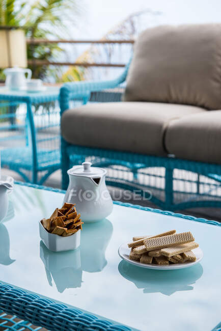 Зверху різноманітні хрусткі вафлі з чайником подаються на плетеному столі зі скляним верхом біля зручного дивана на терасі тропічного готелю — стокове фото