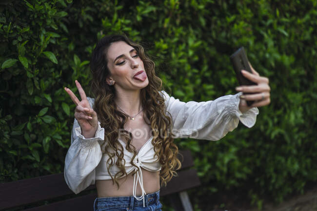 Juguetona joven hembra con el pelo largo rizado mostrando la lengua y v signo mientras toma selfie en el teléfono inteligente en el exuberante parque - foto de stock