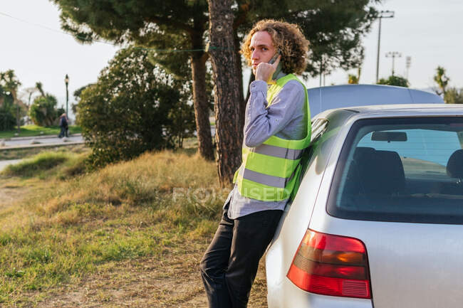 Side view of male driver in safety vest standing near auto with open hood and doing phone call for assistance while getting troubles during trip in countryside — Fotografia de Stock