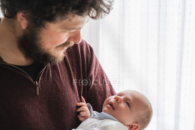 Взрослый бородатый отец с вьющимися волосами, обнимающий милого маленького ребенка, смотрящего друг на друга в доме — стоковое фото