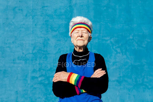 Coole ältere Frau mit Stirnband und Armbändern, die mit verschränkten Armen auf blauem Hintergrund steht — Stockfoto