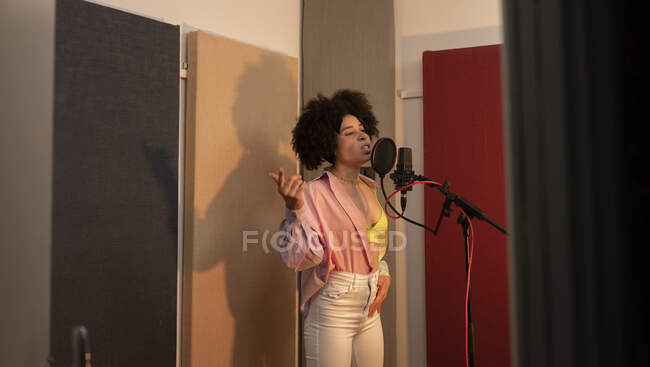 Черная певица исполняет песню против микрофона с поп-фильтром, стоя с рукой на бедре и глядя вперед в звукозаписывающей студии — стоковое фото