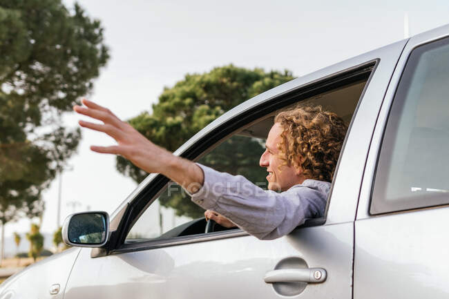 Side view of cheerful young haired male keeping hand out of car window while enjoying summer journey in nature — Stock Photo