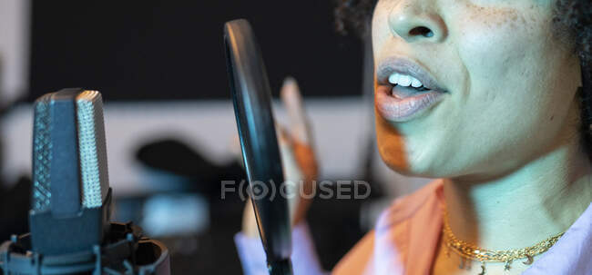 Cantante femenina negra interpretando canción contra micrófono con filtro pop mientras está de pie con la mano en la cadera y mirando hacia adelante en el estudio de sonido - foto de stock