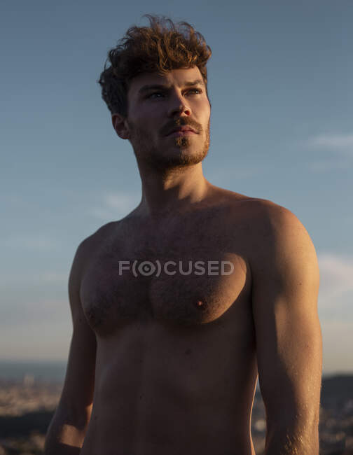 Uomo senza maglietta con sei pack abs e barba guardando lontano contro le montagne sotto il cielo nuvoloso in serata — Foto stock