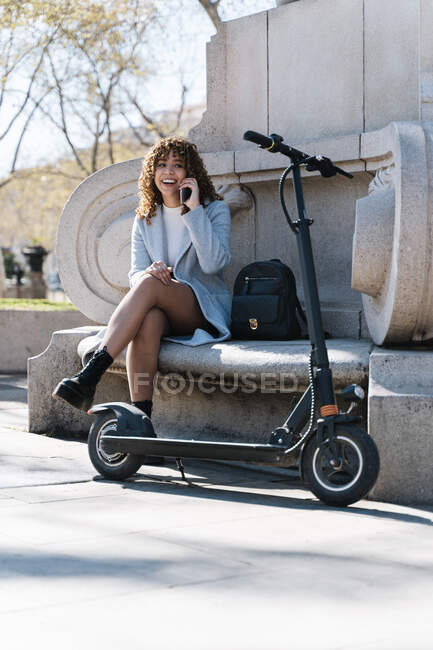 Полное тело молодая весёлая африканка в синем пальто разговаривает по телефону, сидя на каменной скамейке в весеннем парке рядом со скутером — стоковое фото