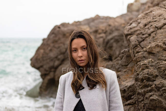 Charmante friedliche Frau im Mantel steht am Sandstrand und blickt in die Kamera — Stockfoto