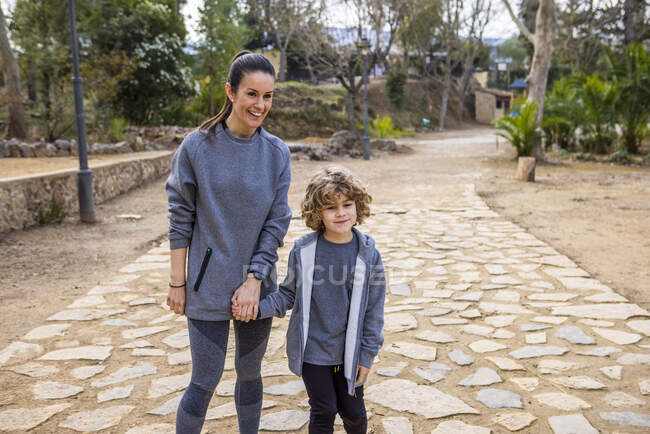 Мать в спортивной одежде держит мальчика за руку, прогуливаясь по дорожке и разговаривая против деревьев глядя на дорогу — стоковое фото