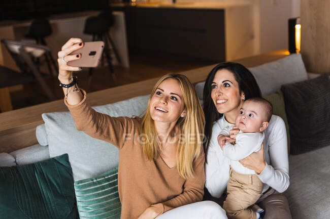 High angle of delighted LGBT family with adorable toddler sitting on couch and taking self shot on smartphone at home — Stock Photo