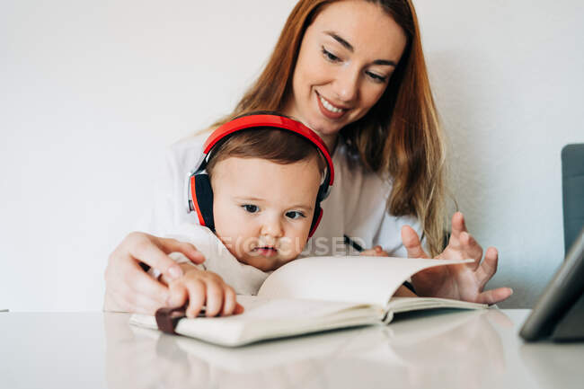 Positive young mother with adorable baby in headphones reading diary while sitting together at desk in light room — Stock Photo