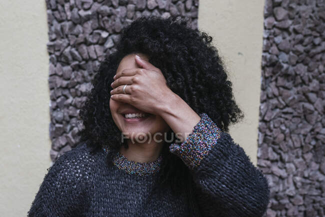 Delighted ethnic female with Afro hairstyle standing on street and covering her eyes with her hand — Foto stock