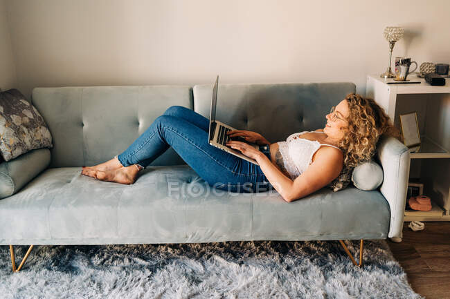 Side view of young self employed woman in casual clothes and eyeglasses lying on comfortable sofa and working remotely on laptop — Stock Photo