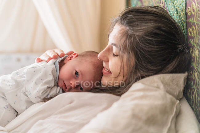 Vista lateral da mãe adulta abraçando gentilmente encantadora criança olhando para a câmera na casa de luz — Fotografia de Stock