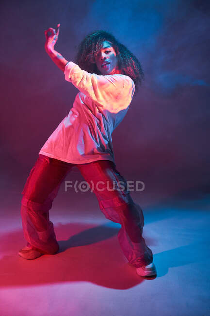Pleine longueur ajustement jeune danseuse afro-américaine en tenue informelle lâche regardant la caméra tout en dansant en studio sombre dans les néons — Photo de stock