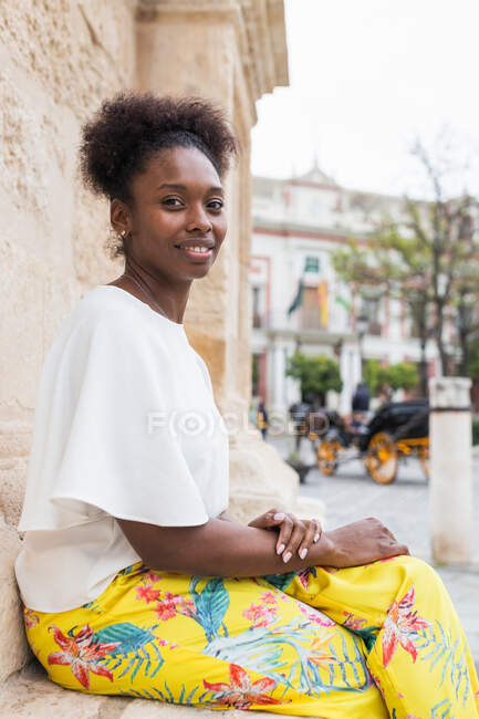 Vista laterale sorridente donna afroamericana in abito alla moda seduta con le gambe incrociate contro il muro e guardando la fotocamera nel soleggiato giardino estivo — Foto stock