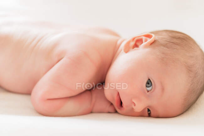 Vue latérale de mignon petit enfant regardant la caméra tout en étant couché sur le textile sur fond blanc — Photo de stock