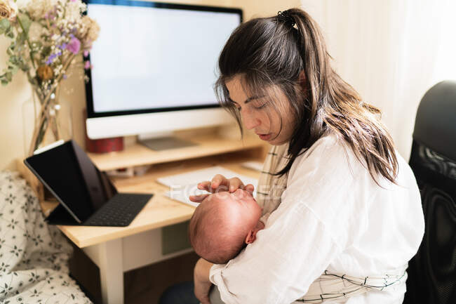 Взрослая мать сидит за столом, работает за настольным компьютером и делает заметки в блокноте, держа маленького ребенка за столом в дневное время — стоковое фото
