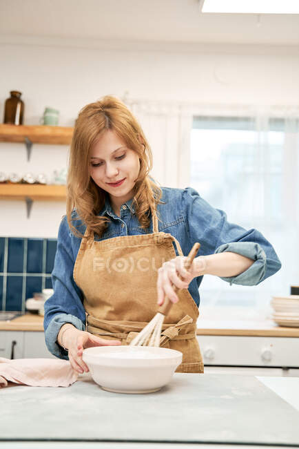 Contenu jeune femme dans tablier fouettant crème sucrée tout en cuisinant à table dans la maison légère — Photo de stock