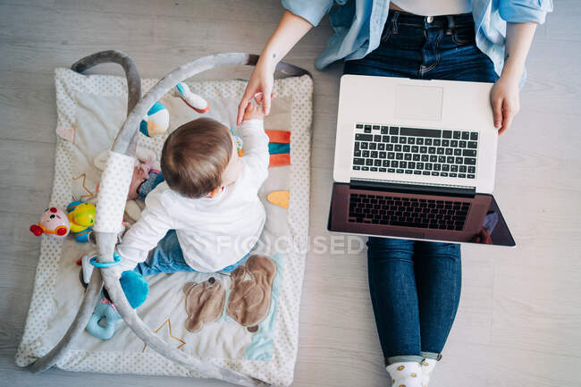 Top view anonymous mother wearing casual outfit sitting on floor with netbook on laps and outstretching hand to cute little baby playing with toys on floor — Stock Photo