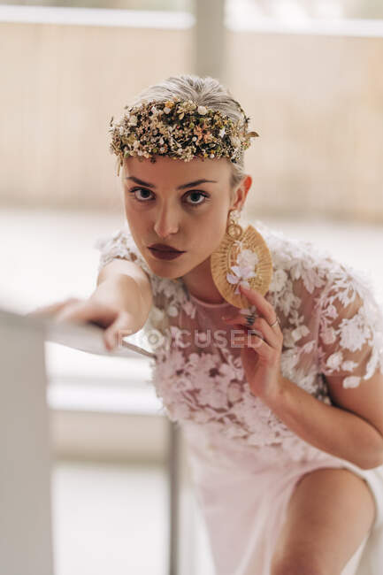 Young female in stylish bohemian white bridal dress and high heeled boots with ornamental wreath and earrings standing on stairway and looking at camera — Photo de stock