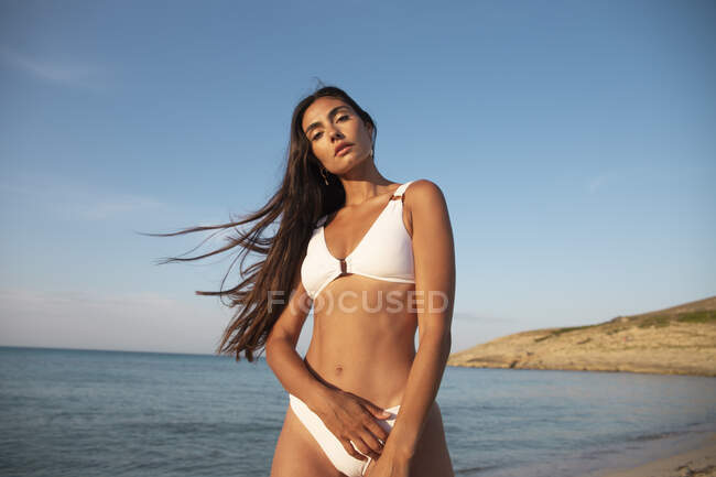 Молодая красивая женщина в купальниках смотрит в камеру, стоя на песчаном берегу океана под облачным голубым небом — стоковое фото
