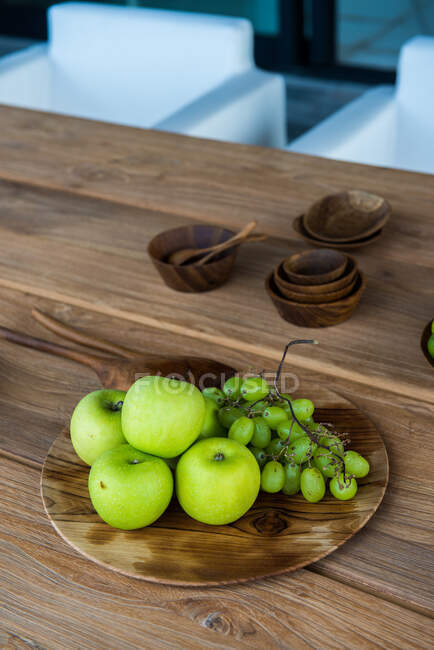 From above of fresh ripe green apples with grapes placed on wooden tray near plate of limes and various traditional bowls served on table in sunlight — Foto stock