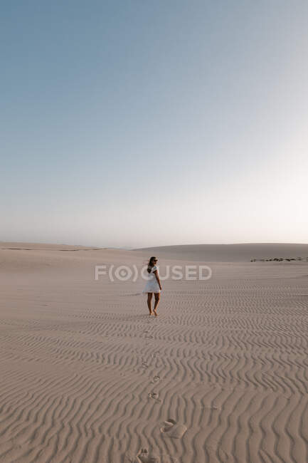 Vista lateral turista feminina em vestido branco passeando na areia com nervuras sob o céu claro — Fotografia de Stock