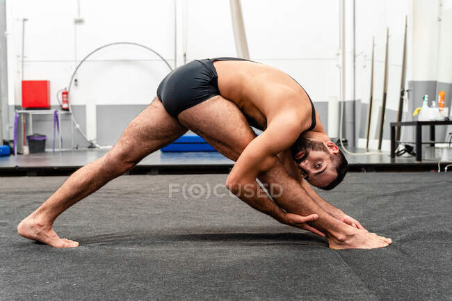 Hombre flexible en forma de cuerpo completo en pantalones cortos deportivos haciendo Triangle Pose y mirando a la cámara mientras practica yoga en el moderno gimnasio - foto de stock