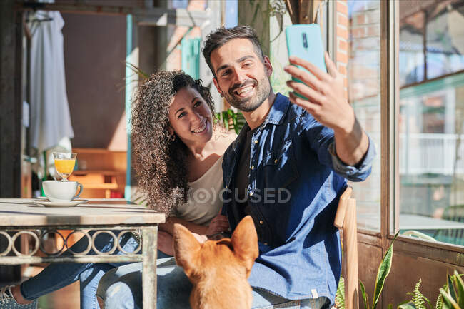 Улыбающийся этнический мужчина с подружкой фотографируют себя по телефону за столом с напитками против собаки в ресторане — стоковое фото