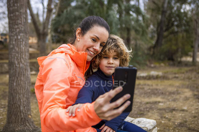 Весела мама в спортивному одязі, що приймає чарівного хлопчика, беручи собі портрет на мобільний телефон у парку — стокове фото