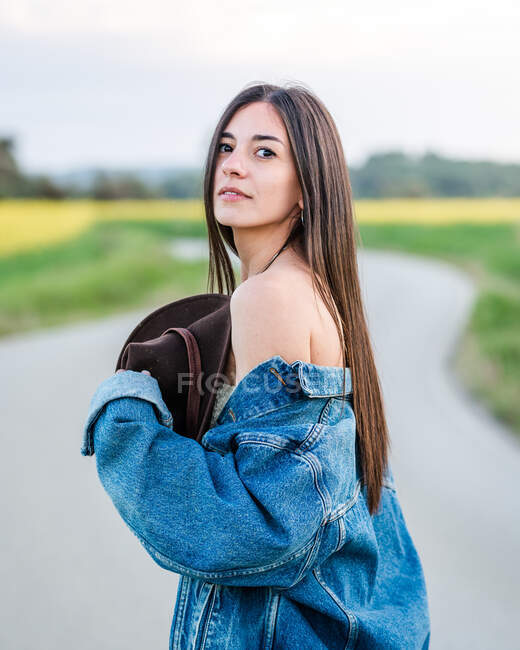 Боковой вид на безмятежную молодую женщину в джинсовой куртке, стоящую на узкой дороге в спокойной летней сельской местности и смотрящую в камеру — стоковое фото