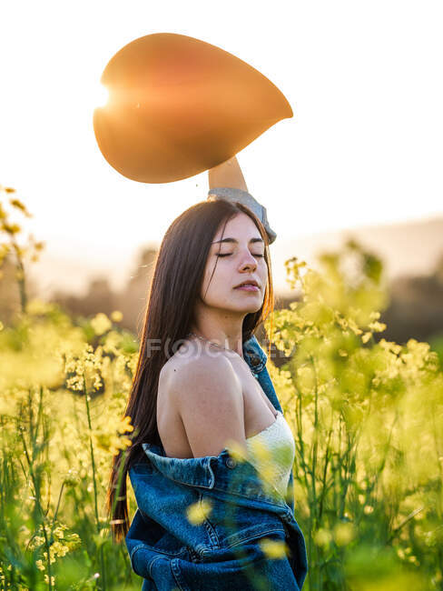 Side view attractive sensitive female in lowered jacket raising hand with hat and standing with eyes closed on sunny blooming rapeseed meadow — Stock Photo