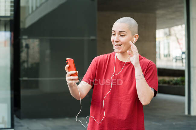 Transgender person in earphones waving hand during video chat on cellphone against building in city — Stock Photo